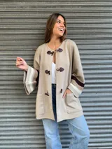 Saco color beige de lana con cierre frontal con broches tipo tanca de cuero. Presenta mangas anchas con detalle de rayas horizontales en marrón y beige.