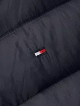 Campera acolchada azul marino con cuello alto, cierre frontal con cremallera y bolsillos laterales con ojal. Presenta detalles distintivos de la marca Tommy Hilfiger, como la cinta con los colores de la marca en el cierre y el logo bordado en el pecho.