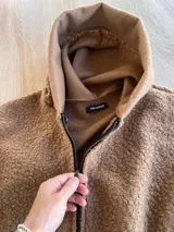 Campera oversize de textura tipo corderito, color marrón, con capucha, cierre frontal completo y dos bolsillos delanteros tipo parche con etiqueta de marca.