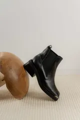 Botas tipo chelsea de cuero color marrón oscuro con acabado brillante. Presentan punta cuadrada con detalle de puntera metálica, elásticos laterales y taco cuadrado bajo.