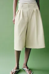 Bermudas de tiro medio color amarillo claro, confeccionadas en mezcla de algodón. Presentan cintura con trabillas, bolsillos laterales ocultos en costura, pliegues delanteros y cierre frontal con cremallera, botón y ganchos metálicos.