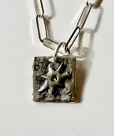 Collar de plata con cadena de eslabones alargados y dije de medalla rectangular con textura rústica, grabado en relieve con forma de estrella y una piedra brillante incrustada en el centro.
