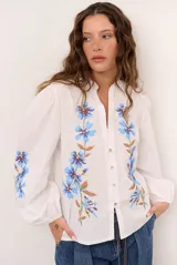 Camisa blanca de manga larga abullonada, con delicados bordados florales en tonos azules y verdes a lo largo de la botonadura frontal y en las mangas. Presenta cuello mao con reborde en contraste y cierre frontal con botones.
