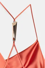 Vestido midi satinado de color naranja tostado, con escote fluido tipo cowl y tirantes finos cruzados en la espalda con detalle de aplicación metálica. Presenta un diseño largo con tajo lateral en la falda.