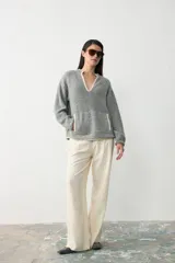 Sweater de tejido bouclé color gris, con cuello en V profundo y bolsillo frontal tipo canguro, ambos con detalles de borde en color crema. Presenta mangas largas y un corte holgado.