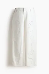 Jean tobillero blanco de corte recto y tiro bajo con detalles de bordado broderie en las piernas. Confeccionado en denim de algodón rígido, cuenta con cinco bolsillos, presillas para cinturón y cierre frontal con botón metálico.