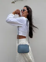 Bandolera pequeña de diseño rectangular en color beige, confeccionada en cuero sintético. Presenta una correa larga ajustable del mismo material y cierre superior con cremallera. Incluye el logo de la marca grabado en la parte frontal.