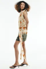 Vestido corto sin mangas de cuello redondo, con estampado tie-dye en tonos beige, naranja y verde oscuro. Presenta un lazo para atar en la cintura y cierre trasero con cremallera oculta.