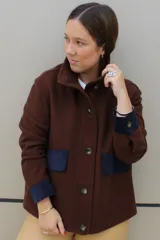 Campera de paño color marrón con detalles en azul marino en los bolsillos y puños. Presenta cuello alto, cierre frontal con botones grandes y corte recto.