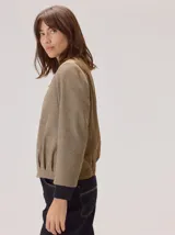 Chaqueta estilo bomber con diseño híbrido, combinando mangas y frente en tejido de lana con patrón de espiga en tonos tierra, y espalda en tejido liso de poliéster color marrón. Presenta puños y ruedo elásticos en color negro, brindando una estructura definida y un estilo contemporáneo.