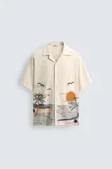 Camisa de manga corta y calce relajado en color beige, con cuello bowling y cierre frontal de botones. Presenta un diseño bordado en la parte inferior con motivos de paisaje tropical que incluyen palmeras, sol y aves.