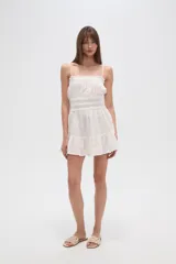 Vestido corto de algodón color blanco, con breteles finos tipo lencero, escote recto con detalle de volado y cintura con elástico fruncido. La falda presenta un diseño holgado con volado en el ruedo.