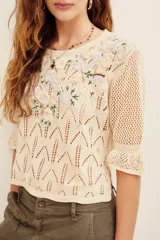 Sweater de tejido calado color beige, con escote en V y mangas cortas abullonadas. Presenta un delicado bordado floral multicolor en la parte frontal superior.