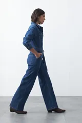 Enterito de jean negro con silueta recta y piernas amplias. Presenta cierre frontal con botones, bolsillos en el pecho y laterales, y cinturón del mismo tejido para ajustar la cintura. De manga larga con puños abotonados.