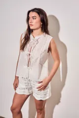 Blusa sin mangas de algodón color blanco off white, con cuello redondo y cierre frontal mediante cordones para atar. Presenta delicados detalles de bordados tonales e inserciones de encaje vertical en el frente.