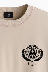 Remera color beige de punto pesado de algodón, de corte holgado, cuello redondo ribeteado y hombros caídos. Presenta estampados en negro en el pecho: el número "33" a la izquierda y un emblema circular con estrellas a la derecha.