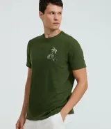 Remera verde militar de algodón texturizado, corte regular, cuello redondo y mangas cortas. Estampa tropical de una casa y palmeras en el pecho.