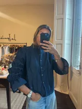 Camisa de denim azul oscuro con cuello mao, cierre frontal de botones metálicos y mangas tres cuartos con diseño fruncido y volados en los puños.