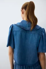 Blusa corta de denim liviano con mangas cortas abullonadas, cuello Peter Pan con volados y cierre frontal con tres tiras para atar.