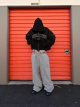 Pantalón deportivo gris de corte ancho y holgado, con cordón de ajuste en la cintura y un estampado de camuflaje sutil en la parte superior, visible bajo la campera.