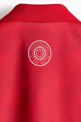 Camiseta de futbol de manga corta de la marca Lotto, en color rojo con un sutil estampado geométrico. Cuenta con cuello tipo polo abierto con escote en V y un calce holgado. Las mangas presentan una franja blanca lateral decorada con el logo de la marca repetido. El logotipo de Lotto destaca en blanco en el centro del pecho.