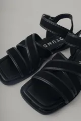 Sandalia plana de cuero negro, con diseño minimalista de tiras finas cruzadas sobre el empeine y una tira ajustable con hebilla metálica en el tobillo.