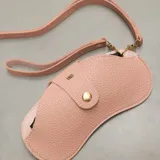 Estuche para lentes de diseño ergonómico y minimalista, confeccionado en cuero sintético con textura granulada. Cuenta con cierre magnético frontal, costuras a la vista en color contrastante y una correa de mano extraíble.
