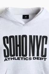 Canguro de felpa gris melange, con capucha de doble capa, bolsillo tipo canguro y estampado frontal con la leyenda 'SOHO NYC ATHLETICS DEPT' en color negro. Presenta hombros caídos y puños y bajo de punto acanalado.