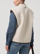 Chaleco de polar tipo corderito en color beige, con cierre frontal y detalles de ribetes en color marrón. Presenta bolsillos laterales y un diseño sin mangas.