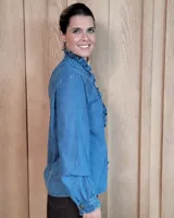 Camisa de denim con volados verticales a lo largo de la botonadura frontal y cuello alto. Presenta mangas largas con puños elásticos y detalle de volados en los mismos.