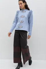 Sweater de lana color celeste, con cuello redondo, mangas largas y volados decorativos en los hombros. Presenta bordados florales artesanales en el frente y detalles de tejido calado vertical.