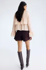 Blusa rosada de tejido liviano, con escote en V rodeado de volados y tiras finas para anudar. Presenta un diseño escalonado con volados anchos en el pecho y en el bajo, y mangas largas acampanadas con volados dobles en los puños.