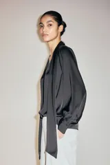 Blusa de gasa negra con cuello alto y lazada frontal, mangas largas abullonadas con puños abotonados y detalle de frunces en los hombros.