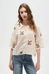 Camisa de algodón color crudo con diseño de paneles unidos por cintas caladas. Presenta bordados florales en cada panel, cuello camisero, cierre frontal con botones y mangas tres cuartos.