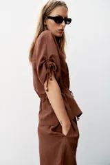 Chaqueta estilo safari de color marrón, con escote en pico, manga corta con cordones ajustables con abalorios y bolsillos delanteros de plastrón. Cuenta con cordón ajustable en la cintura y cierre frontal con botones.