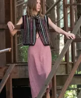 Chaleco tejido a mano en crochet de lana merino color negro, con diseño sin mangas, cuello mao y cierre delantero con cordones para atar. Presenta un patrón de rayas verticales con detalles geométricos en tonos fucsia, celeste y blanco.