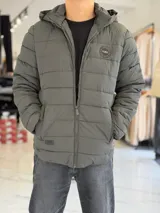 Campera puffer impermeable verde militar con capucha, cierre frontal y bolsillos laterales. Presenta un parche redondo con la inscripción "TURK" en el pecho y un detalle de etiqueta en el dobladillo inferior.