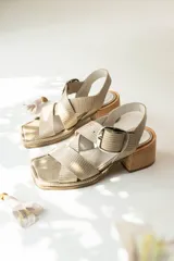 Sandalias de cuero con textura grabada, diseño de punta cuadrada y tiras cruzadas en el empeine. Presentan una hebilla metálica grande lateral, talón descubierto con tira ajustable y taco cuadrado de madera de altura media.