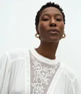 Blusa de viscosa color blanco con cuello alto, mangas largas con puños ajustados y detalles de encaje bordado en el frente y en los puños.