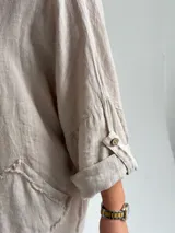 Blusa holgada de lino color beige, con escote en V, mangas tres cuartos con trabilla y bolsillos delanteros aplicados con bordes deshilachados. Cuenta con tiras frontales decorativas con botones de madera.