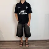 Chomba de tejido de punto color negro, con cuello polo y cierre frontal parcial. Presenta diseño de rayas verticales sutiles en el tejido, detalles de líneas blancas en los hombros y estampado frontal con texto y gráficos en color blanco.