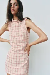 Vestido corto sin mangas de tejido texturizado con diseño de cuadros en tonos crudo y fucsia. Presenta cuello subido, trabillas decorativas con botones dorados en los hombros y cierre lateral con cremallera oculta.