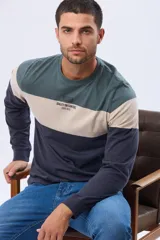 Remera de manga larga con cuello redondo y diseño de rayas horizontales en azul verdoso, beige y azul marino. Presenta una estampa frontal con texto y un parche decorativo en la manga.