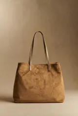 Bolso tipo tote de gran tamaño confeccionado en material sintético con acabado efecto gamuza color tostado. Cuenta con doble asa larga para llevar al hombro y placa metálica pequeña con logo en la parte frontal.