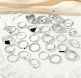 Set de 27 anillos plateados con diferentes diseños: lisos, con forma de serpiente, con corazones, con piedras negras, con forma de mariposa y con eslabones.