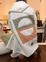 Campera de corderito blanco con capucha, de corte holgado y diseño largo. Presenta un aplique rectangular en la espalda con diseño geométrico de semicírculos en tonos verde y naranja con estampado floral pequeño.