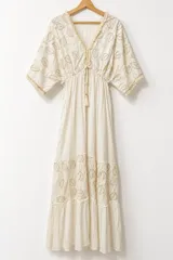Vestido largo color blanco con bordados artesanales en tono beige, escote en V con cordones y borlas, mangas 3/4 estilo murciélago y diseño amplio con volados en la falda.