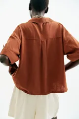 Camisa de lino color celeste claro, con cuello solapa, escote pico, manga corta con vuelta y dos bolsillos de plastrón en el delantero. Cierre frontal con botones.