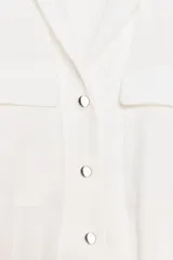 Chaqueta blanca de manga corta con cuello solapa, cierre frontal de botones oscuros y dos bolsillos de plastrón con solapa en el pecho. El bajo es elástico.