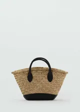 Bolso tipo canasta de paja trenzada color beige con base y asas cortas de cuero sintético negro.